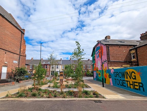 A small urban garden with young trees and plants in the centre of a street. There are red‑brick houses on both sides. A bright mural on a nearby wall shows colourful shapes and the words “Growing Together”. The sky is blue with light clouds.