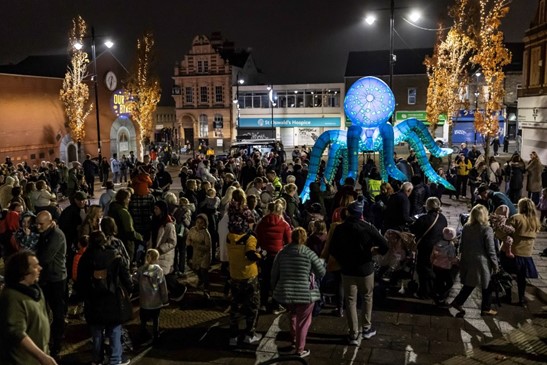 A large crowd gathers in a town square at night for an outdoor event. Decorative trees with warm lights line the space. In the centre stands a large illuminated sculpture shaped like an octopus, glowing blue and green. Buildings and shopfronts surround the square, creating a lively atmosphere.