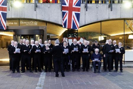 Around 25 men from the Newcastle Male Voice Choir sing outside the Civic Centre. They are all dressed in black trousers, white shirt and black jumpers. One older man is sitting, the rest are all standing.