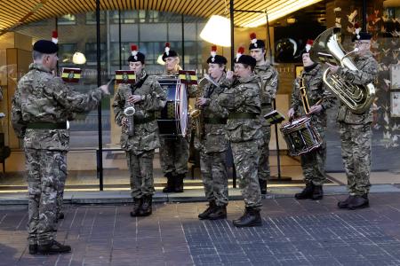 Band of the Royal Regiment of Fusiliers play outside the Civic Centre. They are wearing combat fatigues with their distinctive caps with the red and white hackle. There are two drummers, one trombone player, two saxophones and a flute player and a conductor.
