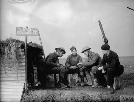 Two British and two French soldiers sit outside a dugout. Thee is a makeshift flag flying over the shelter with Downing street written on it. They are sitting at a table playing cards. One soldier has a gun on his shoulder pointing up towards the sky.