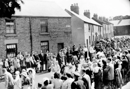 Men, women and children line both sides of a street of terraced houses on VE Day 1945. Children in fancy dress and playing musical instruments form a parade along the middle of the road.