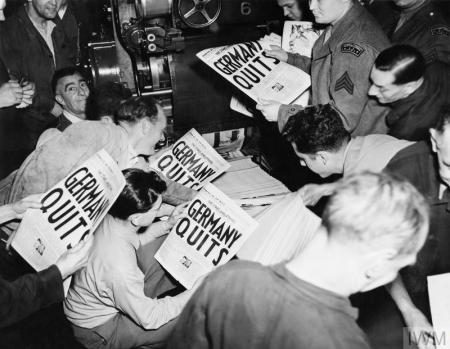 Journalists in a newsroom reading copies of a newspaper with the headline Germany quits. The image is in black and white and was taken in 1945.