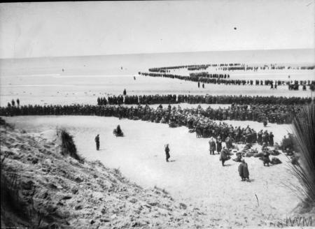 British troops wait to be rescued from a in Dunkirk on a dull and cloudy day. They are wearing uniform and carrying kit bags and light weapons. Despite the bad weather some have waded into the sea while others sit or stand patiently on the beach.