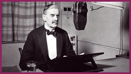 Prime Minister, Neville Chamberlain, is sat at a desk ready to broadcast to the nation. He is wearing a black suit, white shirt and black bow tie. Hanging down in front of him is a large radio microphone.