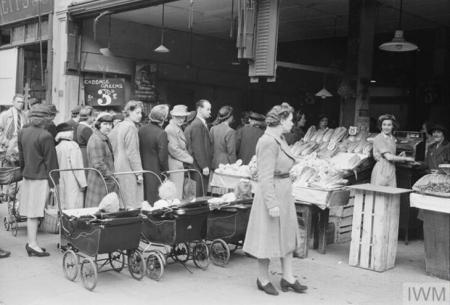 A queue of around 20 women and two or three men outside a greencrocers. There are three prams with babies in at the side of the queue.