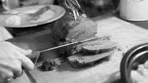 A cooked stuffed heart on a chopping board. A hand can be seen slicing the heart with a meat knife. The roasting dish is next to the chopping board and contains the roasted vegetables.