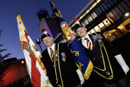 Two veterans from the Royal Fusiliers stand outside the Civic Centre. They are wearing their veterans uniforms and caps with the distinctive red and white hackle. They each hold a raised Standard.