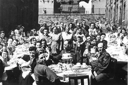 VE Day Street Party in Newcastle. Children and adults sitting at rows of tables enjoying the day. Some men are in uniform. All are smiling at the camera. There is food and drink on the tables including cakes on cake stands and china tea cups and saucers.