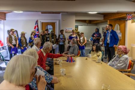 Four older women and three older men sit around a long table in the middle of a room at Byker lodge. There is a Union Jack hat on the table and some cups of tea and glasses of wine.Six girls and four boys wearing army hats are standing at the far end of the table. They are reading stories from World War 2. Behind them is a Union Flag and the room is decorated with bunting. Three members of staff stand to one side of the table watching the activities.