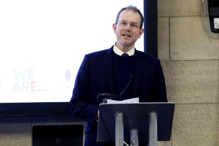 Rabbi in a black suit and white shirt stands at the podium in the Banqueting Hall at the Civic Centre. There is a large welcome screen behind him