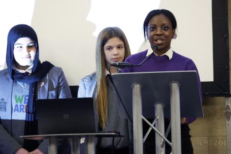Three female students at the memorial day event. The girl at the front is Black. She is wearing a royal blue uniform jumper with a white shirt underneath. Her hair is tied back. Slightly behind to her right is a white girl. She is wearing a light blue jacker and has long, light brown hair that hangs straight to her waist. To her right is a Muslim girl. She is wearing a light grey blazer and a navy headscarf