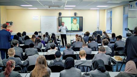 A group of secondary school students sitting at desks in a classroom. They are wearing grey school blazers and are facing the front of the classroom. Smajo Beso is standing at the front of the class and talking to the students. He is wearing a light grey top and black trousers. He has a beard and wears glasses. Behind him is a large screen which has a picture of a young boy on it.