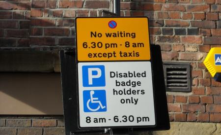 The shared use sign designated for taxis and disabled badges. The first part of the sign is yellow with the no waiting symbol and states ‘No waiting 6.30pm – 8am except taxis’. Underneath is the standard disabled sign which has a picture of the disabled wheelchair symbol and states ‘Disabled badge holders only 8am – 6.30pm’