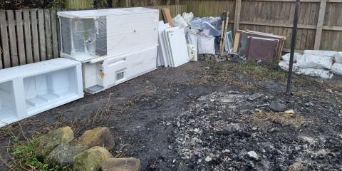 Partially dismantled fridges stacked against a wooden fence, surrounding scorched earth