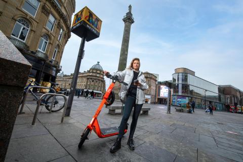 A woman is standing with an orange e-scooter in the centre of Newcastle next to a Metro station and with Grey's Monument in the background.