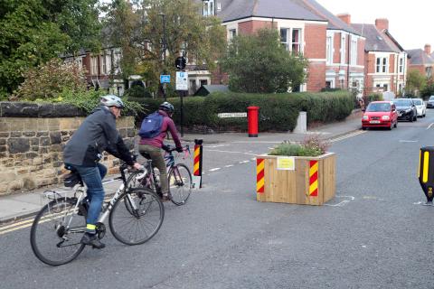 Two people on bikes ride along a street, with houses on the left,  that has been closed to vehicles using bollards and wooden planters. 