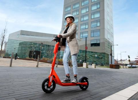 Photo showing a young woman using an orange e-scooter on a clear road with a tall building in the background. She is wearing light coloured clothing and a white helmet.