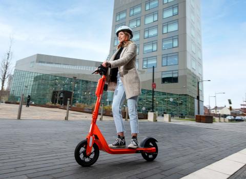 Photos shows a young woman riding an orange e-scooter with a tall building in the background.