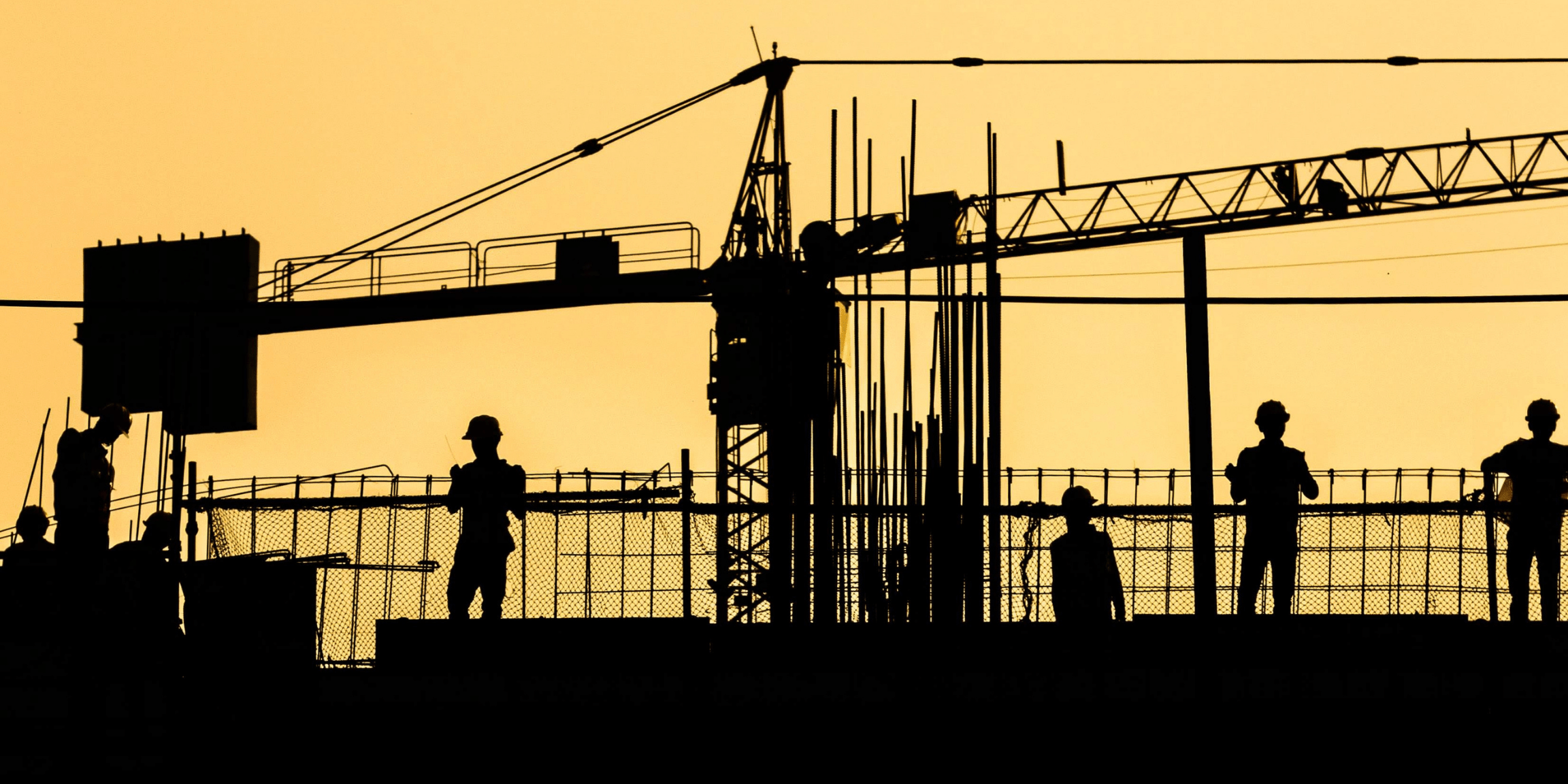 Construction workers and a tower crane silhouetted against an orange sky