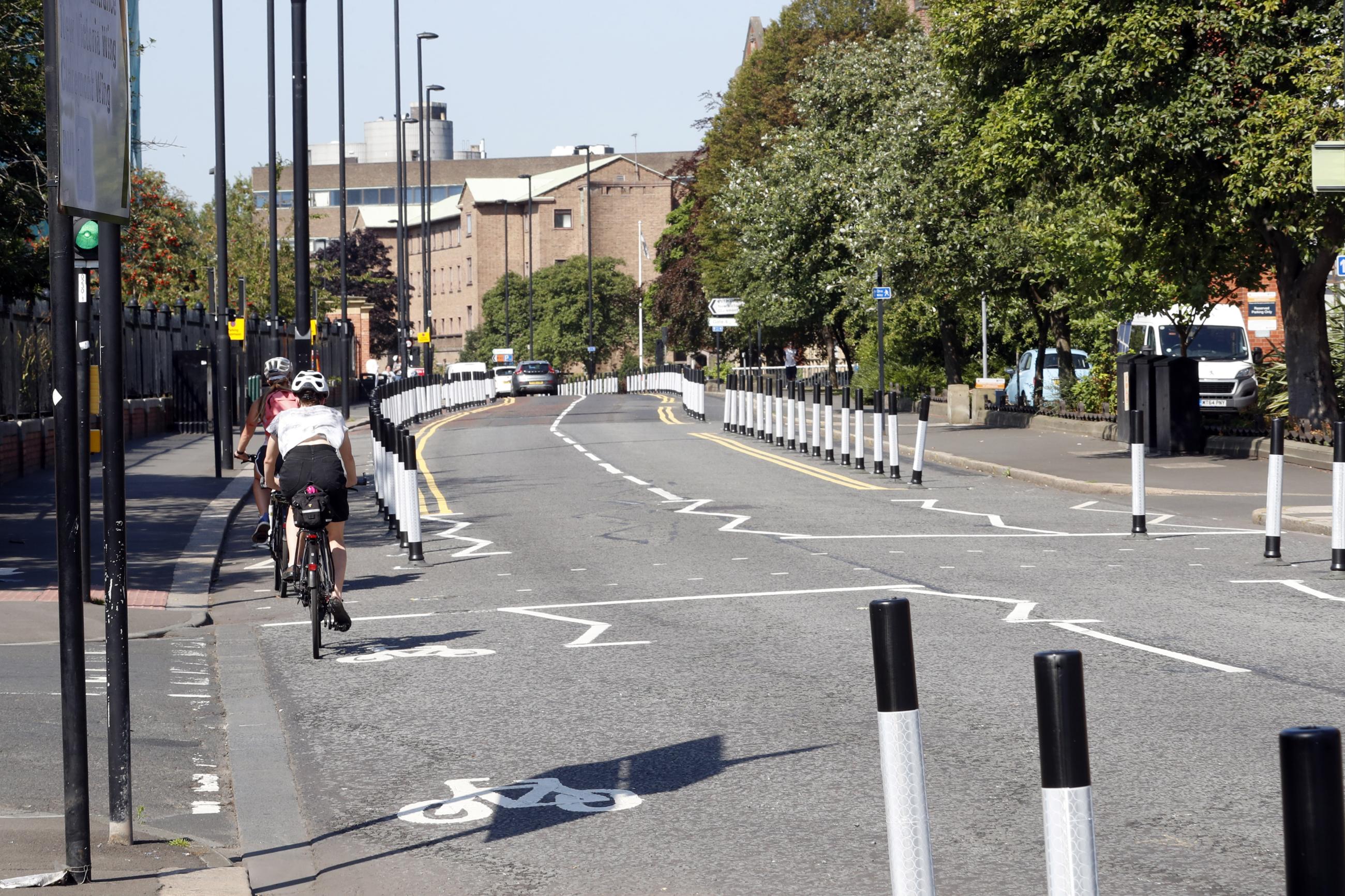 Photo shows a stretch of road with two cyclists travelling away from the camera along a temporary cycle lane marked out with black and white posts