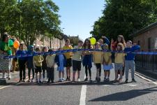 Children from Hotspur Primary School lined up across the road with a large ribbon that one child is cutting to mark the official opening of their School Streets scheme.
